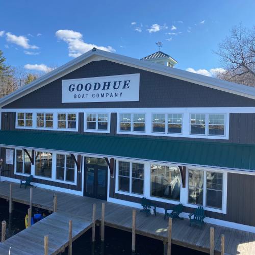 A large building with "Goodhue Boat Company" sign, featuring green roof and multiple windows, by a dock on a sunny day.