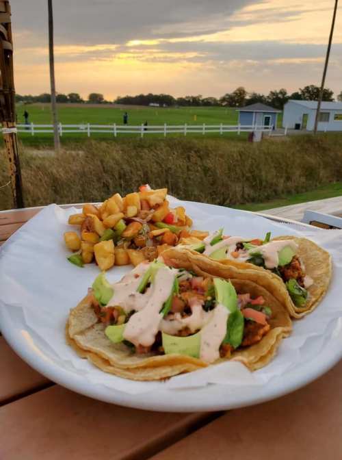 Two tacos with toppings and sauce on a plate, accompanied by seasoned potatoes, with a sunset and fields in the background.