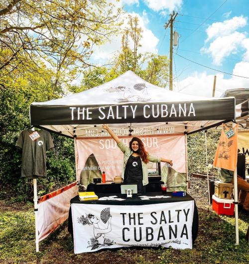 A vendor booth with a sign reading "The Salty Cubana," featuring merchandise and a smiling person behind the table.