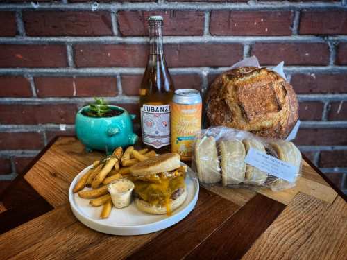A plate with a cheeseburger, fries, a bottle of drink, a can, and a loaf of bread on a wooden table.