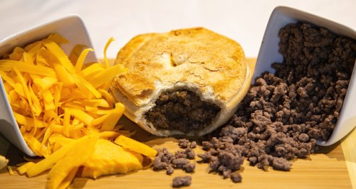 A savory meat pie with a bite taken out, surrounded by shredded cheese and crumbled ground beef on a wooden board.