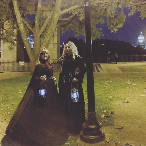 Two women in dark, elegant costumes hold lanterns in a park at night, with a city skyline in the background.