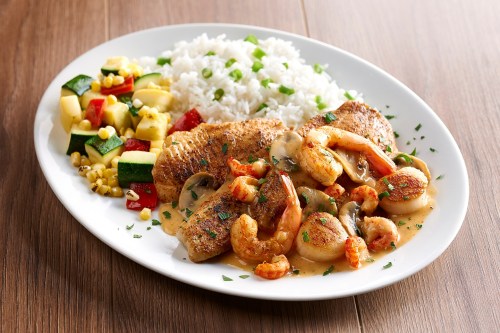 A plate of grilled fish and shrimp in sauce, served with rice and a colorful vegetable medley.