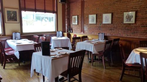 Cozy restaurant interior with wooden floors, brick walls, and neatly set tables with white tablecloths.