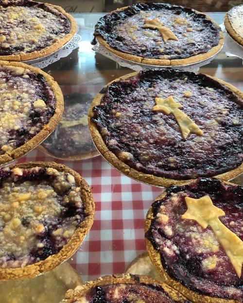 A display of various berry pies with crumbly toppings on a checkered tablecloth.