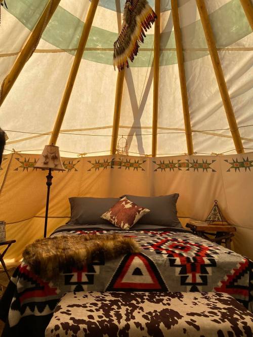 Interior of a cozy teepee with a bed, patterned blankets, a lamp, and decorative elements. Warm, inviting atmosphere.