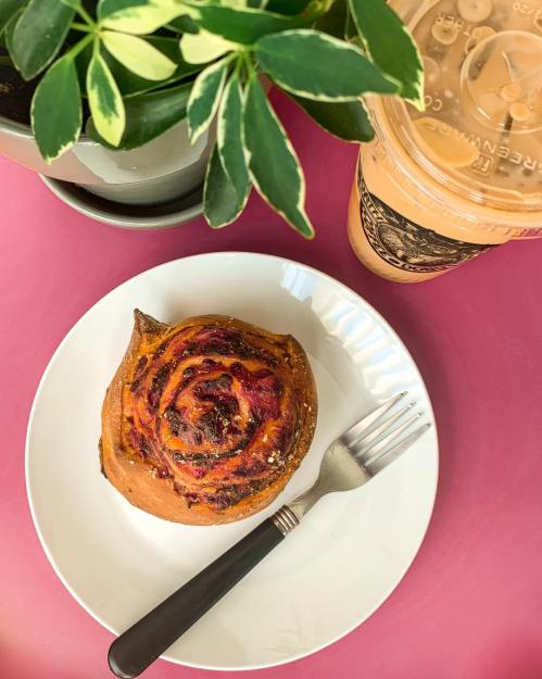 A cinnamon roll on a white plate with a fork, next to a plant and a cup of iced coffee on a pink table.