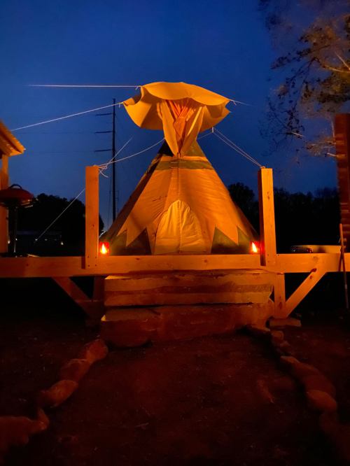 A lit teepee at night, surrounded by wooden structures and a pathway, creating a warm, inviting atmosphere.