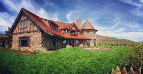 A picturesque stone house with a red roof, surrounded by green grass and mountains under a blue sky with clouds.