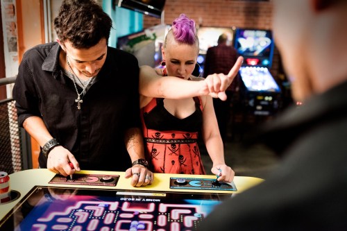 A man and a woman play arcade games, with the woman gesturing playfully while focused on the screen.