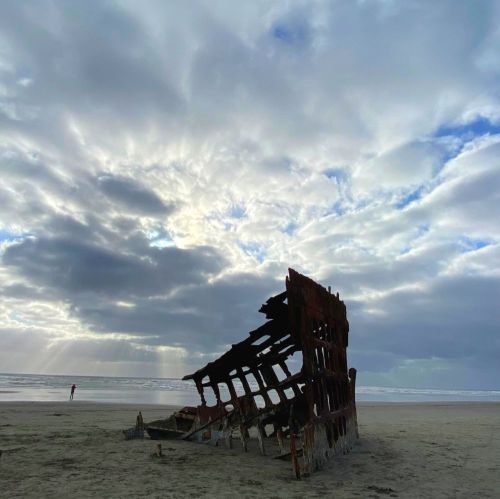 A shipwreck on a sandy beach under a cloudy sky, with a person standing in the distance.