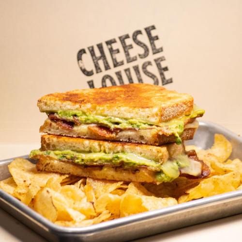 A stacked grilled cheese sandwich with bacon and avocado, served with a side of crispy potato chips.