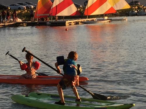 Two children paddle on colorful kayaks in a calm lake, with vibrant sailboats in the background during sunset.