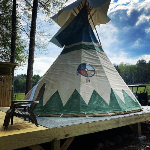 A colorful teepee on a wooden deck surrounded by trees and a cloudy sky. A chair sits nearby.