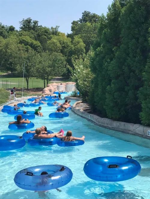 People float on blue inner tubes in a lazy river at a water park, surrounded by greenery and a water slide in the background.