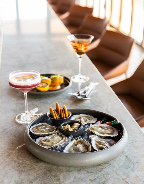A platter of oysters on ice with snacks, accompanied by two cocktails on a marble table.
