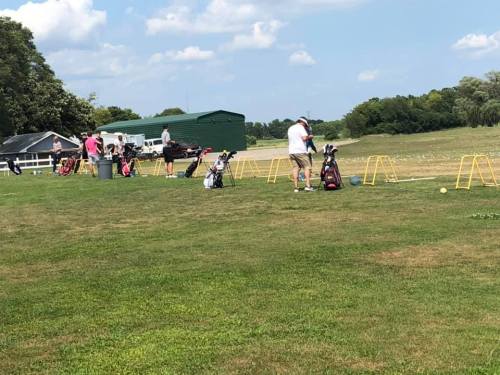 A golf driving range with several people practicing their swings, surrounded by green grass and a blue sky.