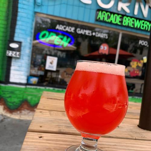 A vibrant red drink in a glass sits on a wooden table, with an arcade brewery in the background.
