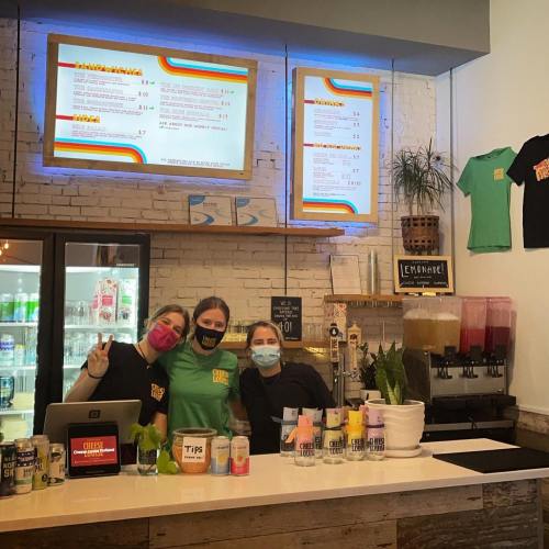 Three staff members wearing masks smile behind a counter with drinks, a menu, and a tip jar in a vibrant café setting.