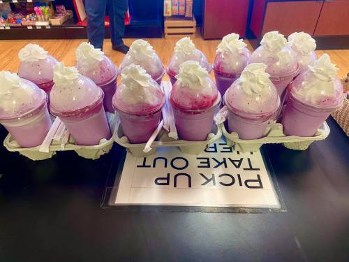 A tray of purple milkshakes topped with whipped cream, placed on a table with a sign that says "Pick Up Take Out."