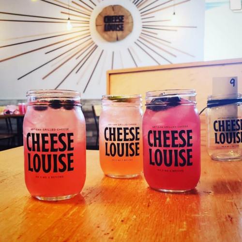 Three drinks in jars with "CHEESE LOUISE" labels on a wooden table, with a sunburst design in the background.