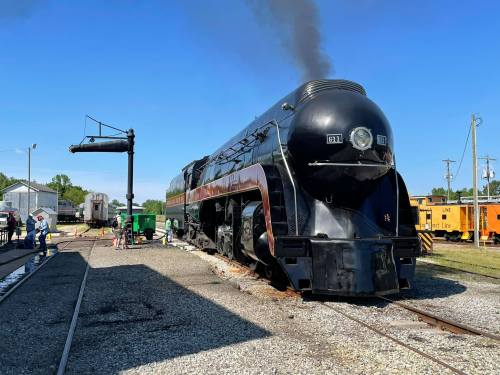 A vintage steam locomotive with smoke billowing, parked at a train station under a clear blue sky.