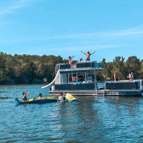 A group of people enjoying a sunny day on a houseboat, with some kayaking nearby in a calm river.