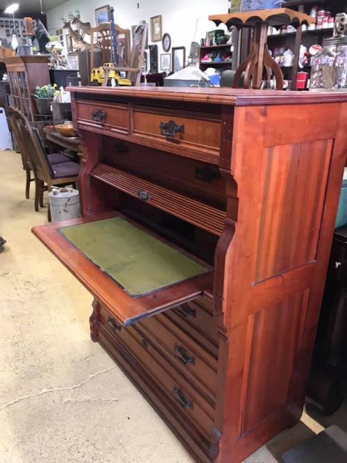 A vintage wooden dresser with multiple drawers and a fold-out writing surface, displayed in an antique shop.