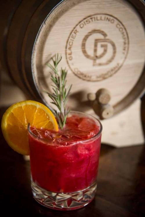 A vibrant red cocktail garnished with an orange slice and rosemary, set against a wooden barrel backdrop.