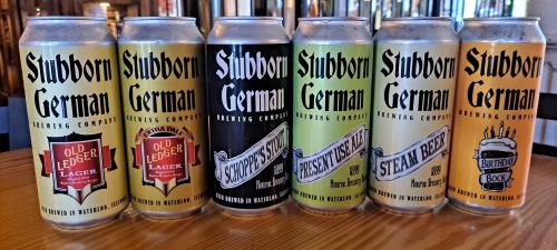 Six cans of Stubborn German Brewing Company beer, featuring various labels: Old Ledger Lager, Schippes Stout, and more.