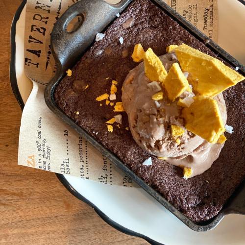 A warm brownie topped with ice cream and crushed toppings, served in a skillet on a decorative plate.