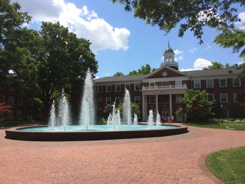 A fountain in front of a brick building with a dome, surrounded by trees and a blue sky with fluffy clouds.
