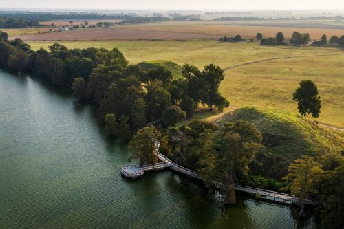 Aerial view of a serene lake surrounded by green trees and a grassy landscape, with a wooden dock extending into the water.