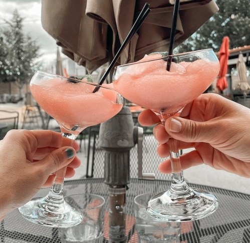 Two hands clink glasses of pink frozen cocktails with black straws, set on a table outdoors.