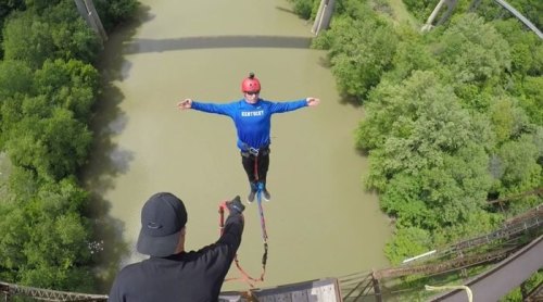 A person in a blue shirt is bungee jumping off a bridge, while another person holds the bungee cord below.