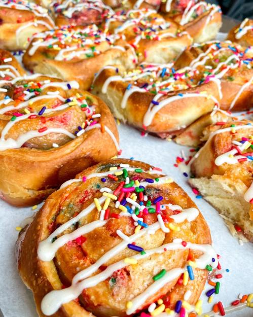 A close-up of colorful cinnamon rolls topped with icing and vibrant sprinkles, arranged on a white surface.