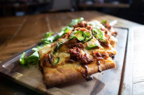 A close-up of a flatbread pizza topped with cheese, jalapeños, and sausage, served on a wooden board with arugula.