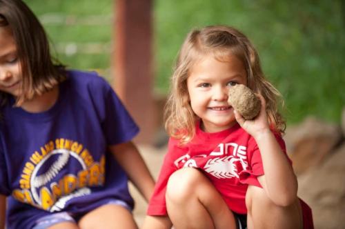 A smiling girl in a red shirt holds a rock, sitting next to another child in a purple shirt outdoors.