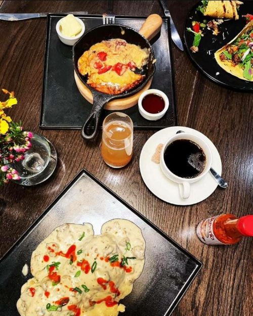 A table set with a skillet dish, pancakes with sauce, a cup of coffee, and a drink, surrounded by flowers.