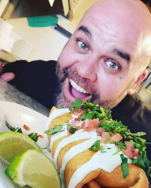 A smiling chef poses with a plate of crispy tacos topped with cilantro, tomatoes, and sour cream, alongside lime wedges.