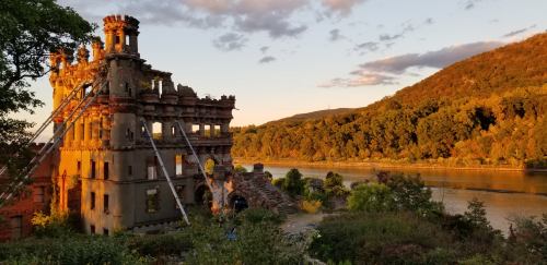 Ruins of a historic building by a river, surrounded by trees and hills, illuminated by a warm sunset.