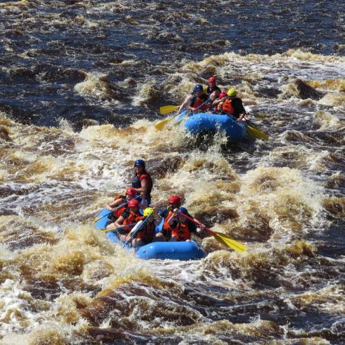 A group of people in blue rafts navigating through turbulent, brown river rapids.