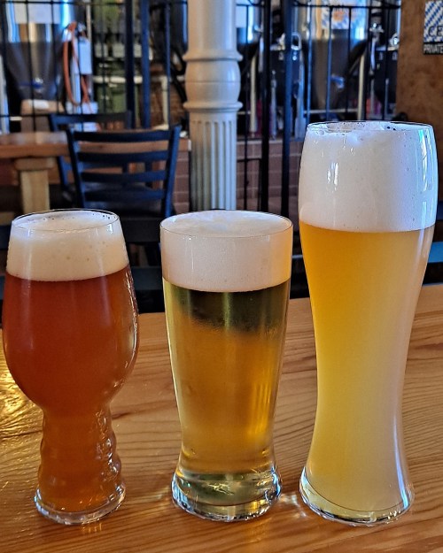 Three glasses of beer in varying sizes and colors, placed on a wooden table in a casual setting.