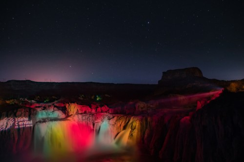 A vibrant waterfall illuminated in colorful lights under a starry night sky, with rocky cliffs in the background.