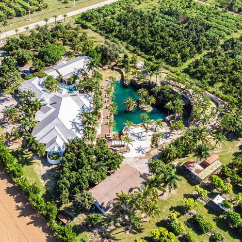 Aerial view of a tropical property featuring a house, pool, and lush greenery surrounding a pond.