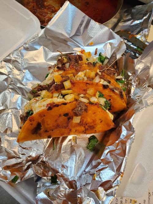 Three tacos filled with meat, onions, and cilantro, served on foil with a side of sauce.