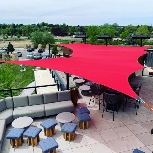 A rooftop patio with red shade sails, modern seating, and a view of greenery and parking in the background.