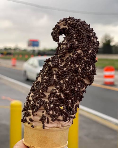 A chocolate ice cream cone topped with a swirl of chocolate sprinkles, held against a cloudy background.