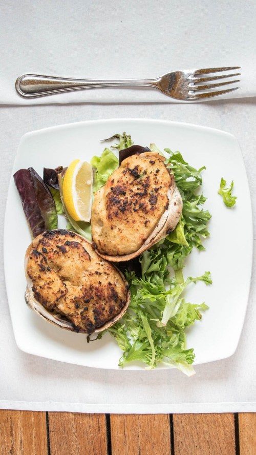 Two stuffed mushrooms on a bed of mixed greens, garnished with a lemon wedge, served on a white plate.