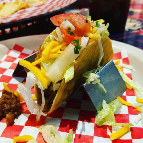 A taco filled with seasoned meat, lettuce, cheese, and tomatoes, served on a plate with red and white checkered paper.